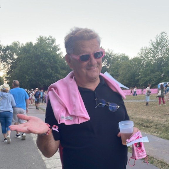 Un homme en lunettes roses, une serviette rose sur les épaules, un verre à la main en plein air